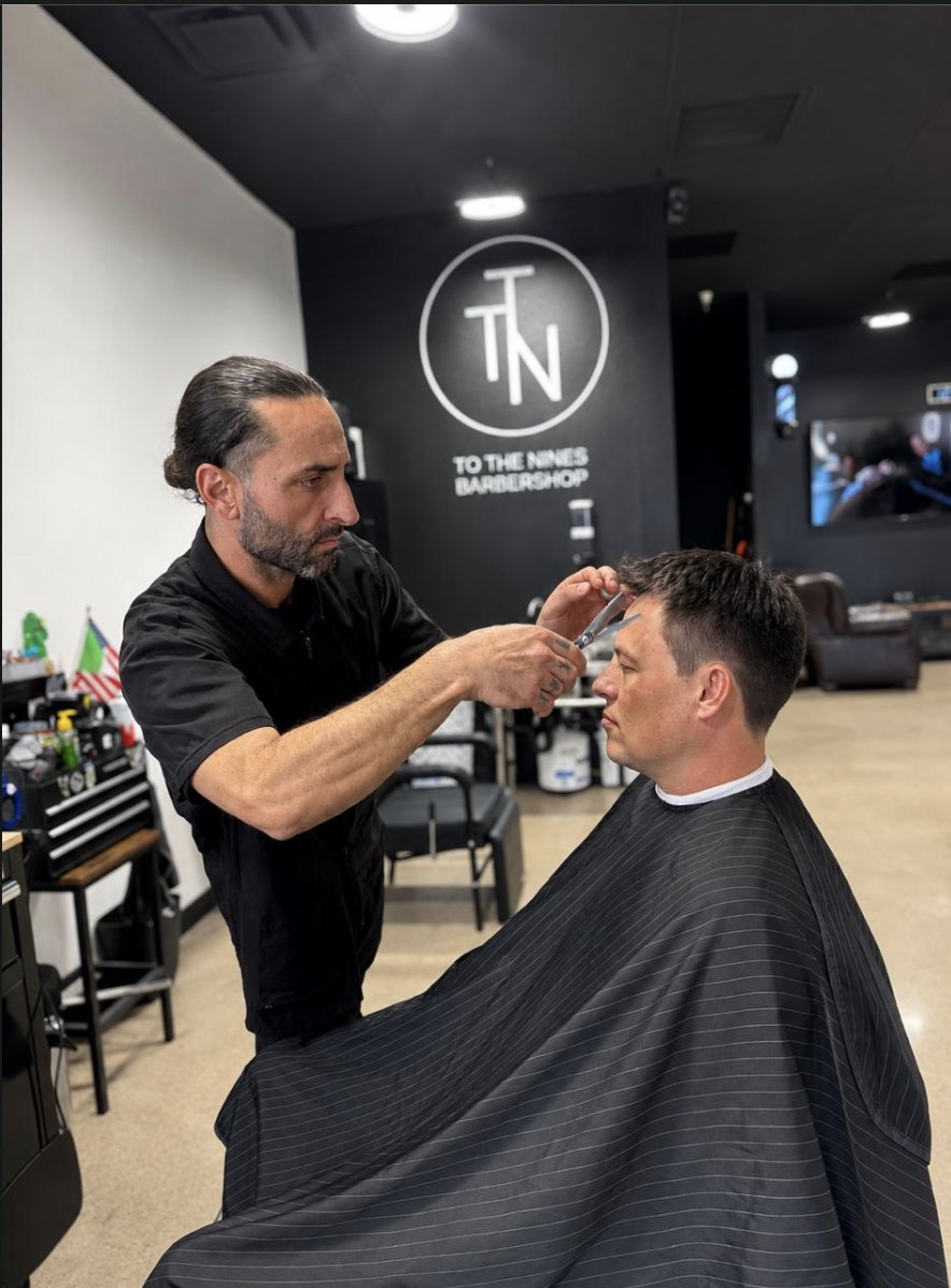 Barber scissor cutting a client's hair under the To the Nines logo wall in Tempe